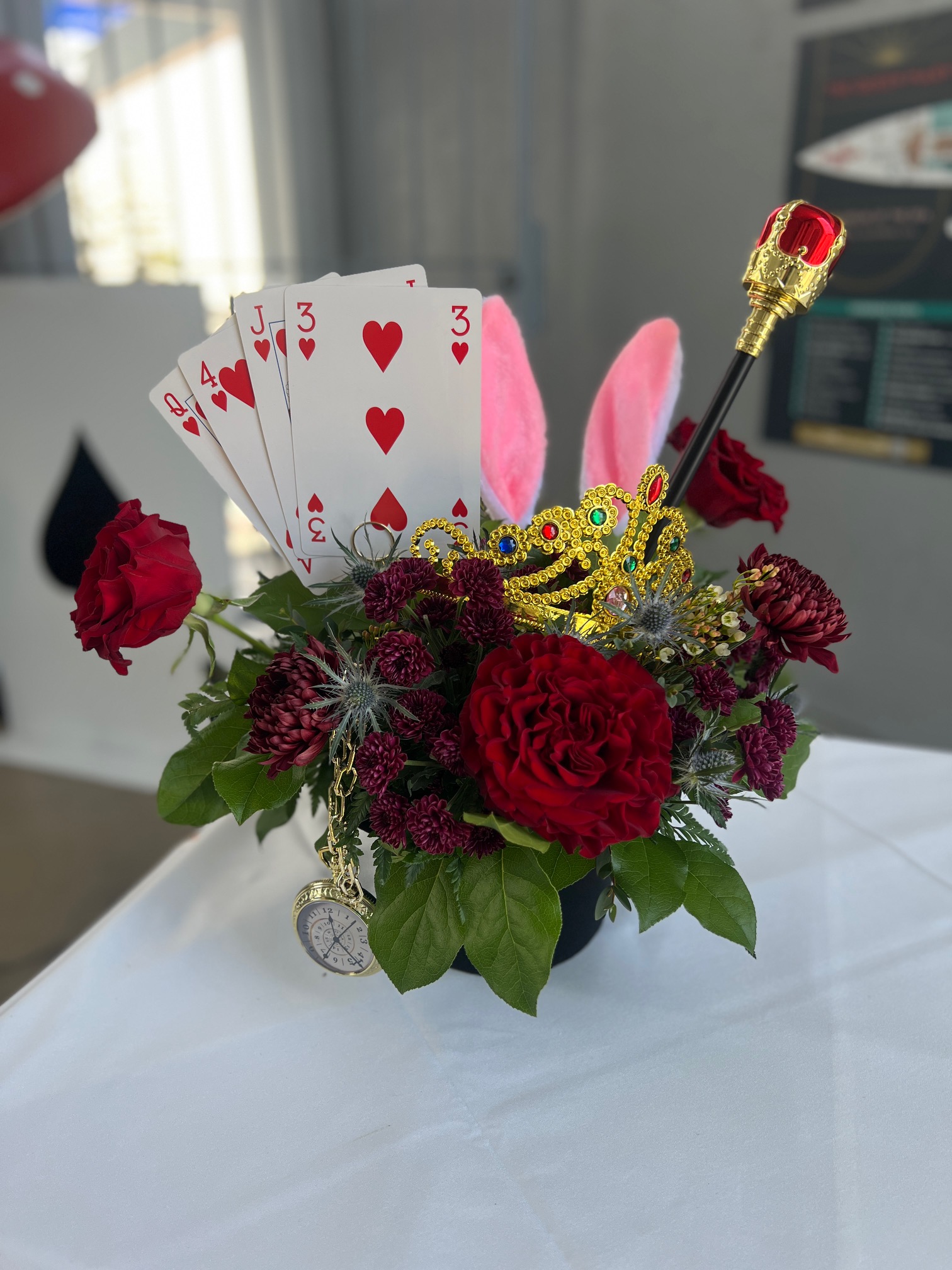 Whimsical red floral centerpiece decorated with playing cards, a jeweled crown, pocket watch, bunny ears, and gold scepter.