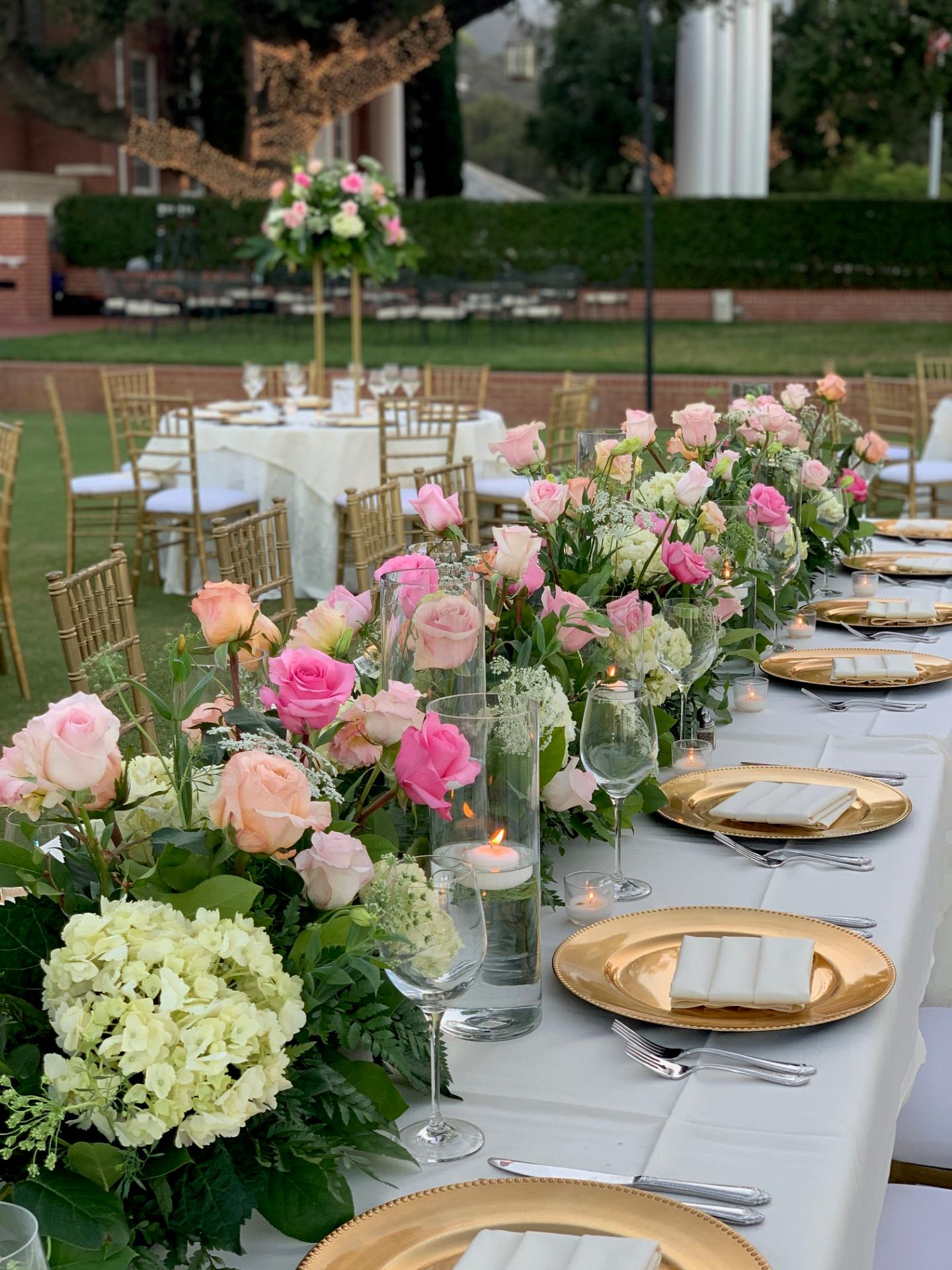 Outdoor reception table decorated with pink and blush roses, white hydrangeas, greenery, floating candles, and gold place settings.