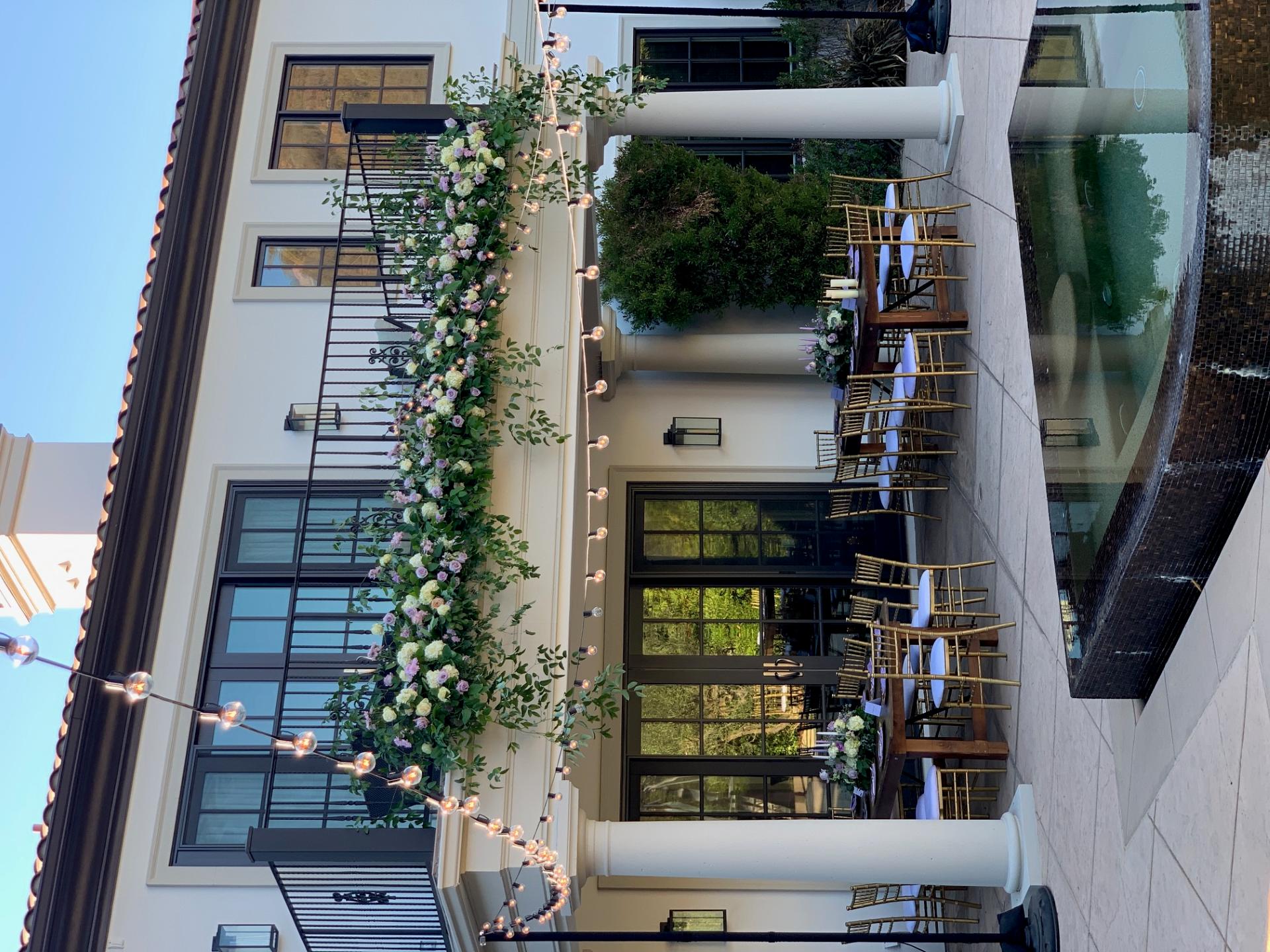 Poolside event setup with floral garlands along a balcony, string lights, wooden dining tables, and gold chairs.
