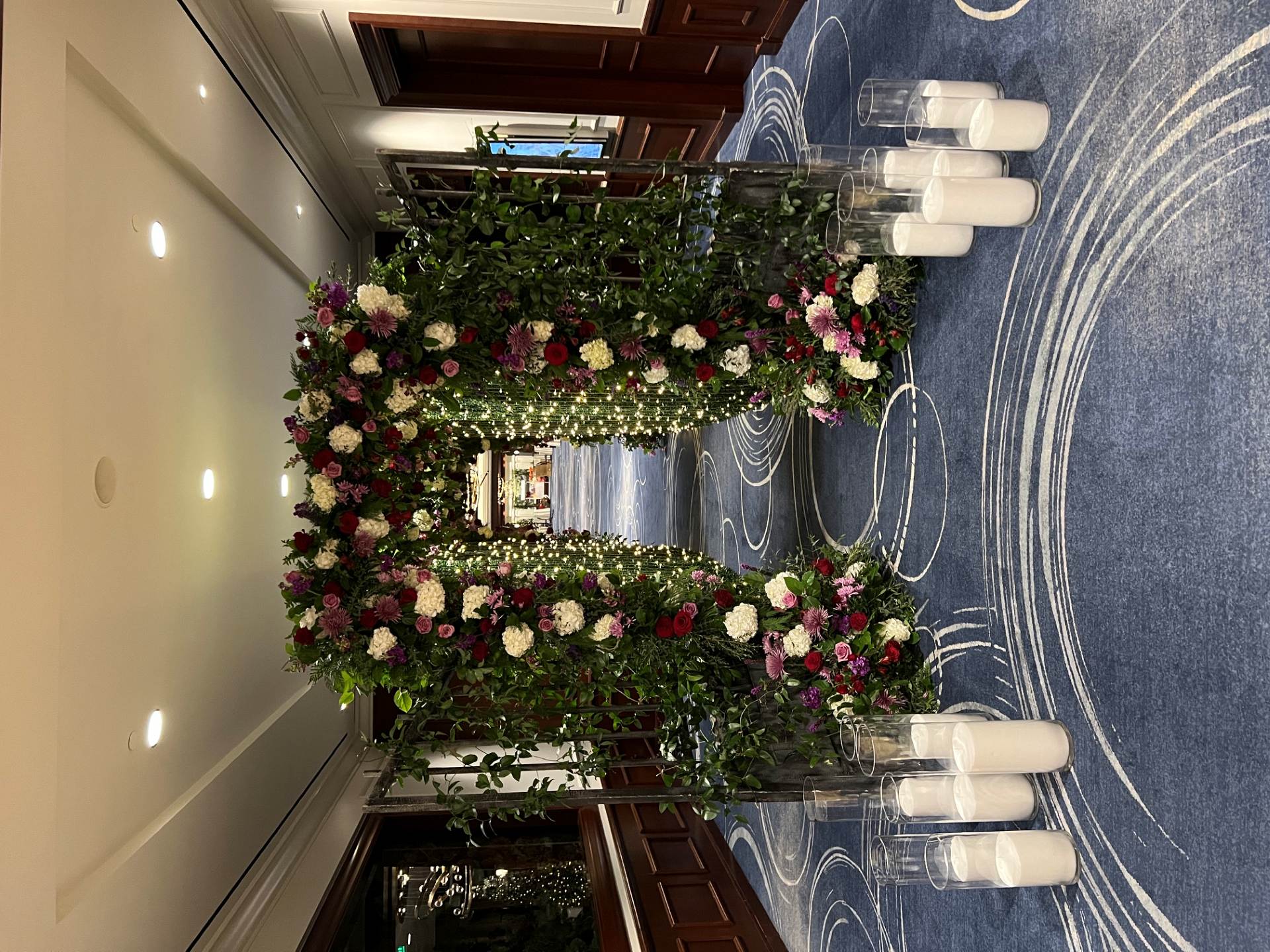 Walk-through floral arch covered in red, pink, and white flowers with lush greenery, string lights, and pillar candles.
