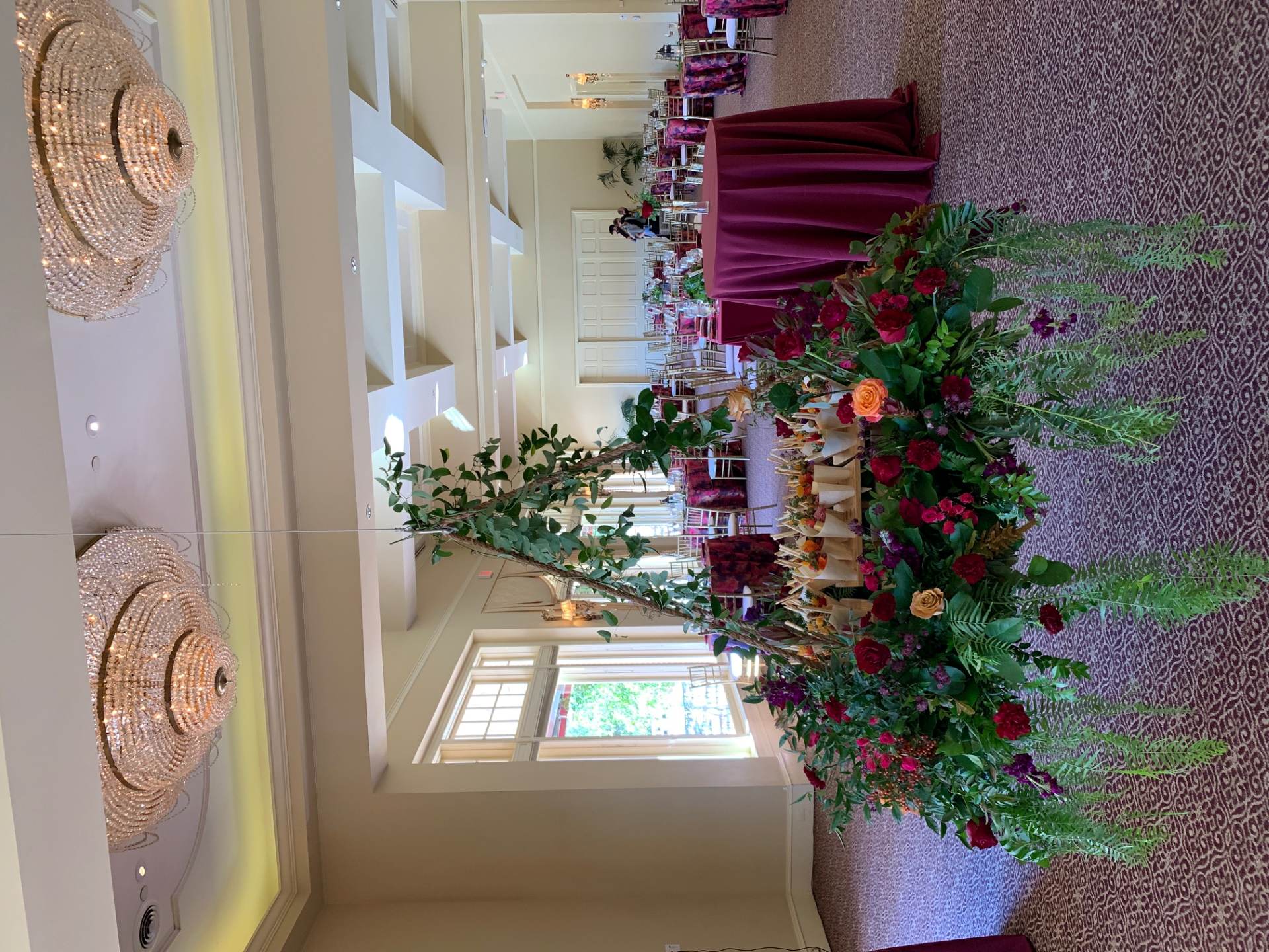 Large suspended floral display with deep red flowers, greenery, and trailing foliage surrounding a central arrangement in a ballroom.