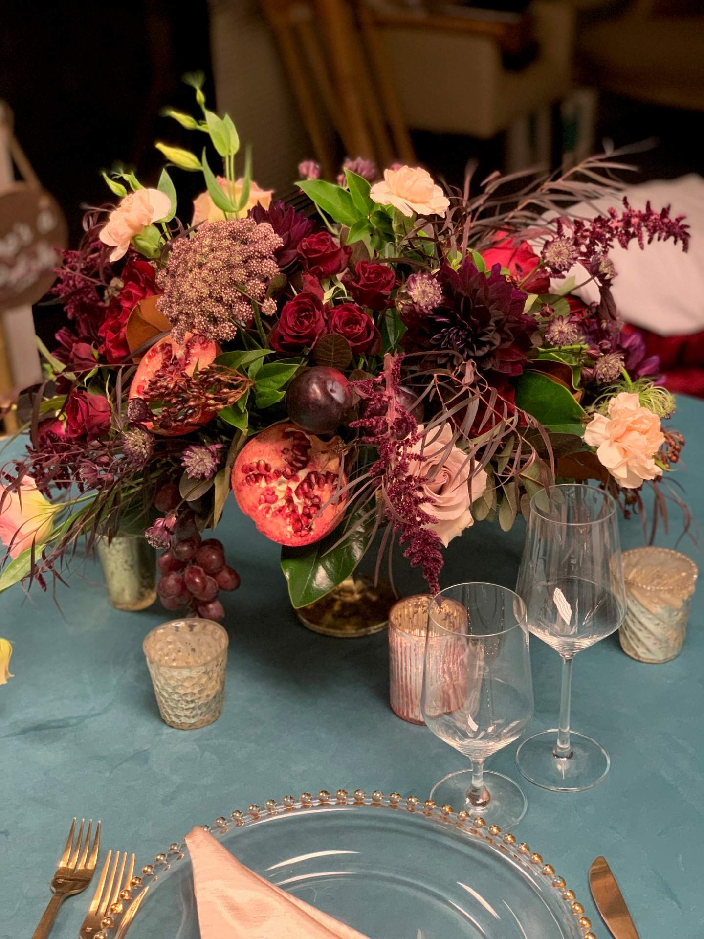 Rich burgundy and peach floral centerpiece with roses, pomegranate, grapes, greenery, and candleholders on a teal table.