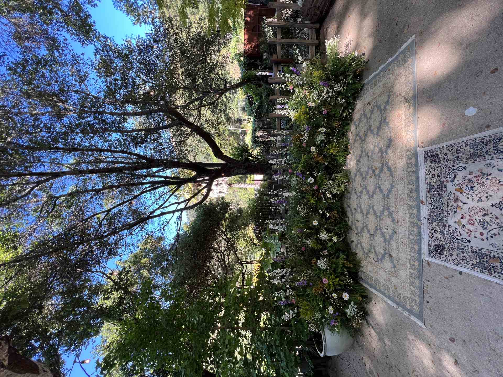 Outdoor garden ceremony area with a long arrangement of white, pink, and purple flowers beneath a large shade tree.