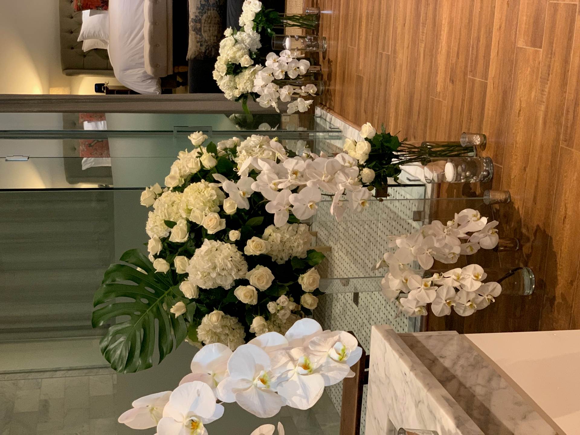 Modern white floral installation with orchids, roses, hydrangeas, tropical leaves, and candles inside a contemporary room.
