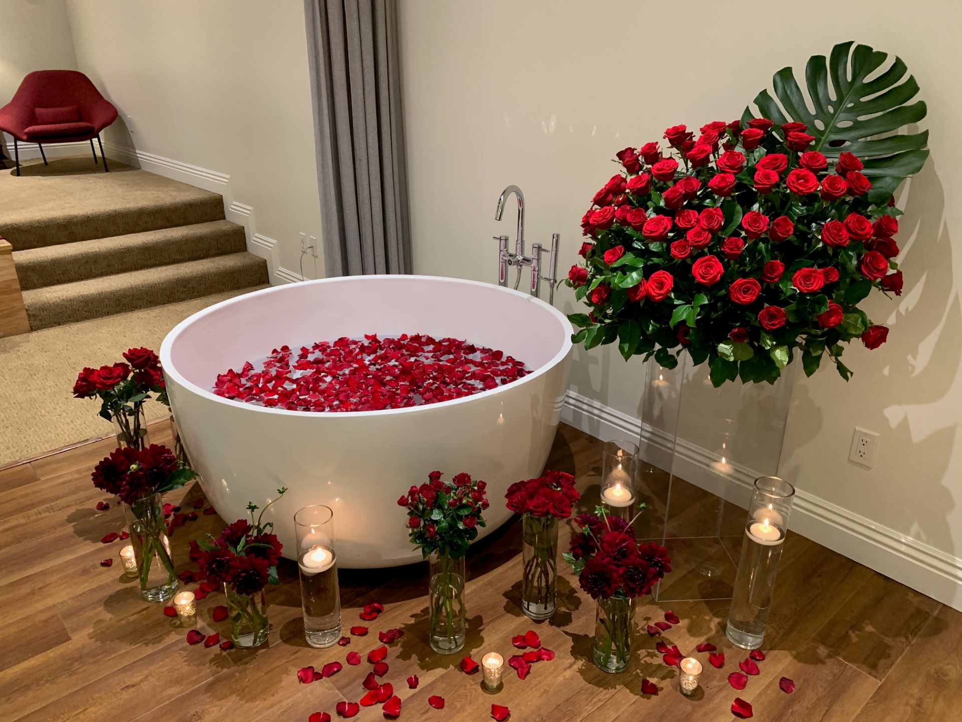 Romantic bathtub setup filled with red rose petals and surrounded by red floral arrangements, candles, and greenery.