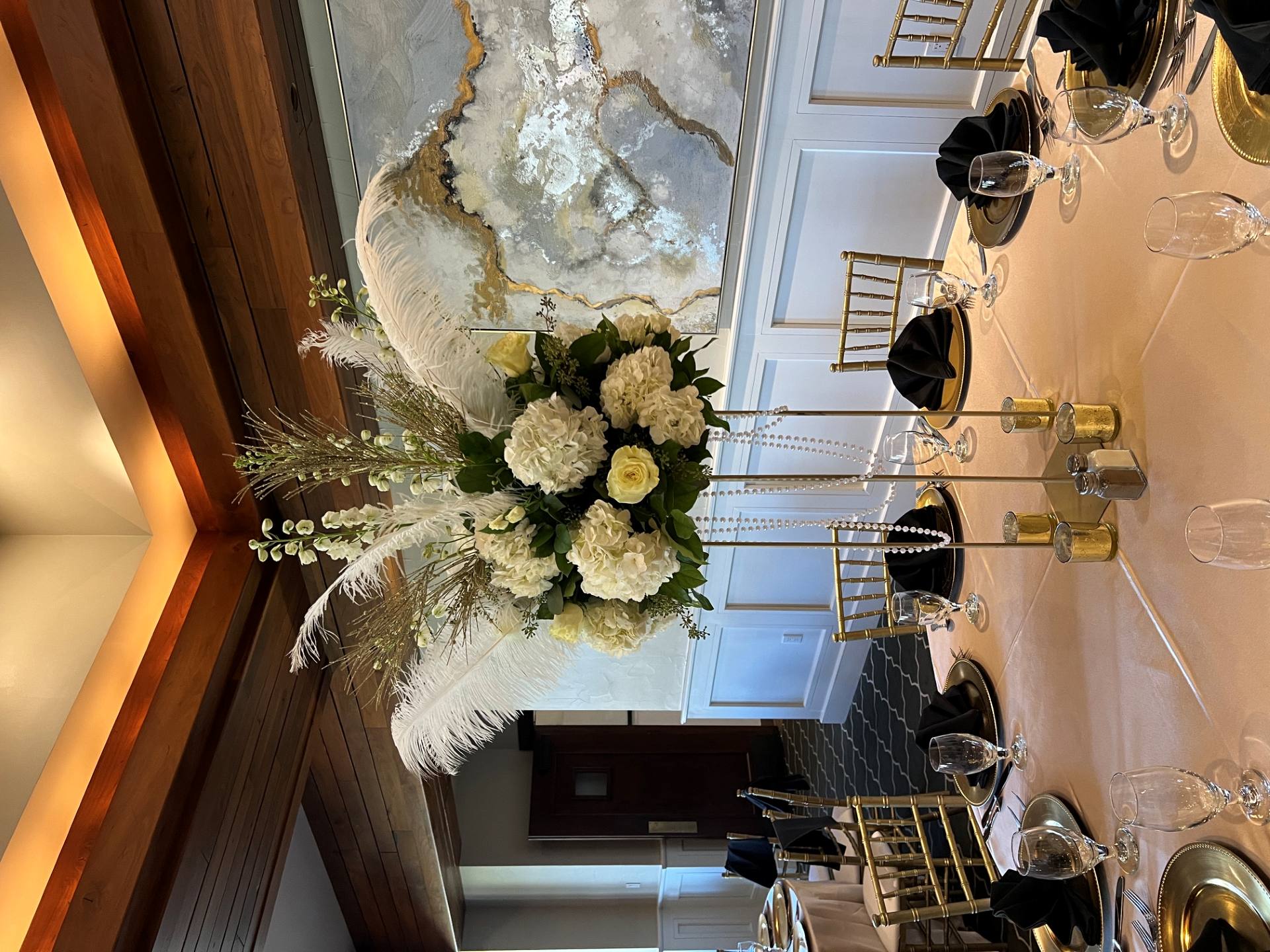 Tall white floral centerpiece with hydrangeas, roses, feathers, greenery, and hanging crystal strands above an elegant dining table.