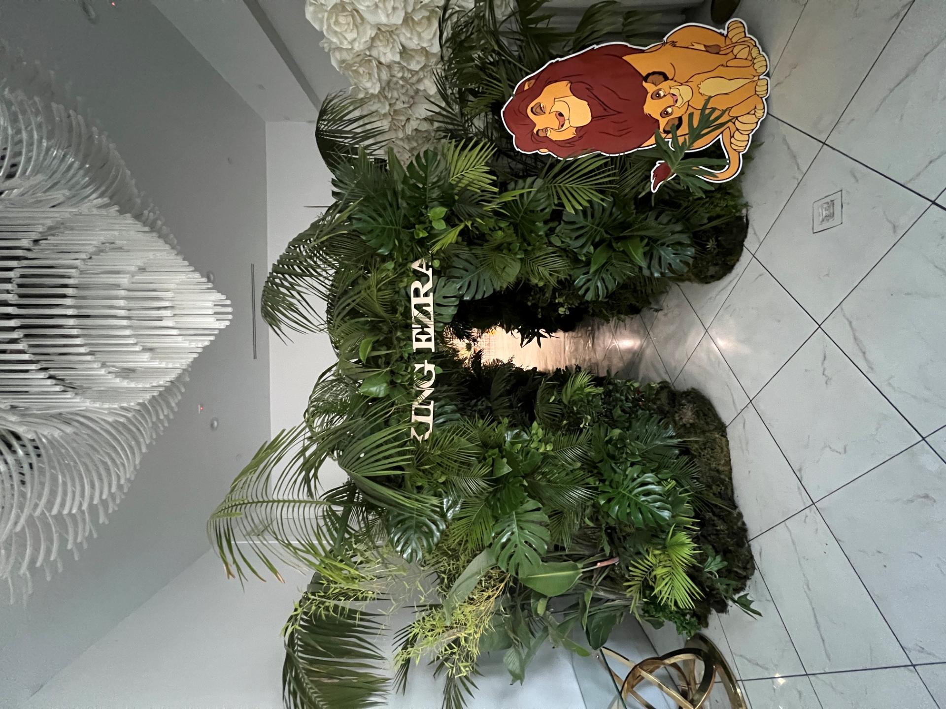 Tropical entrance installation covered in dense palm leaves and greenery with a Lion King themed display beside the walkway.