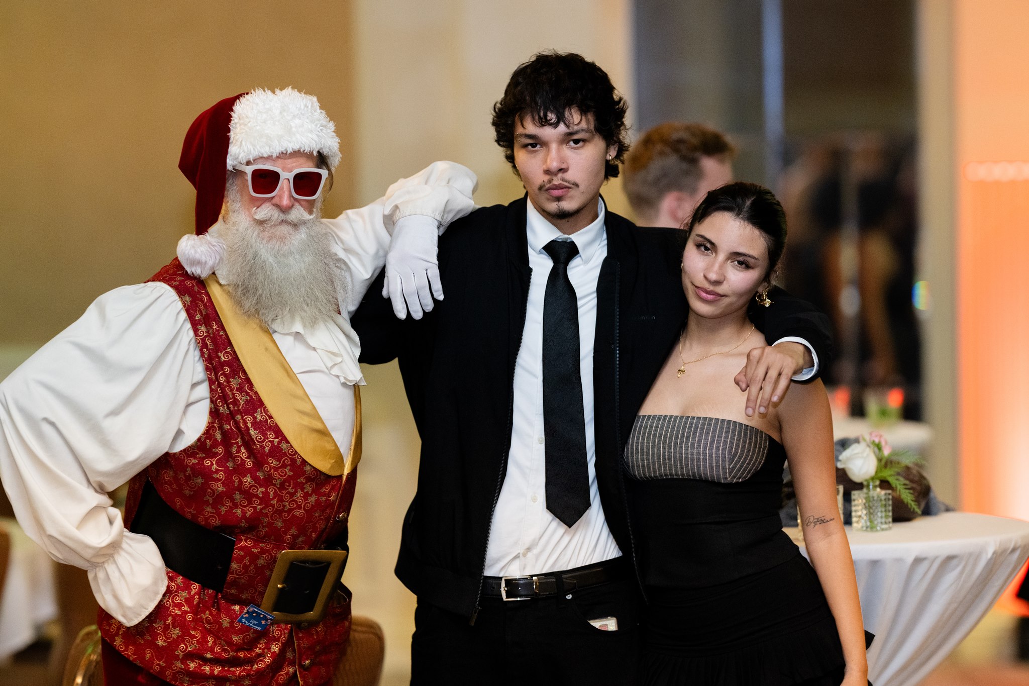 Santa posing with two formally dressed guests at an indoor holiday event.