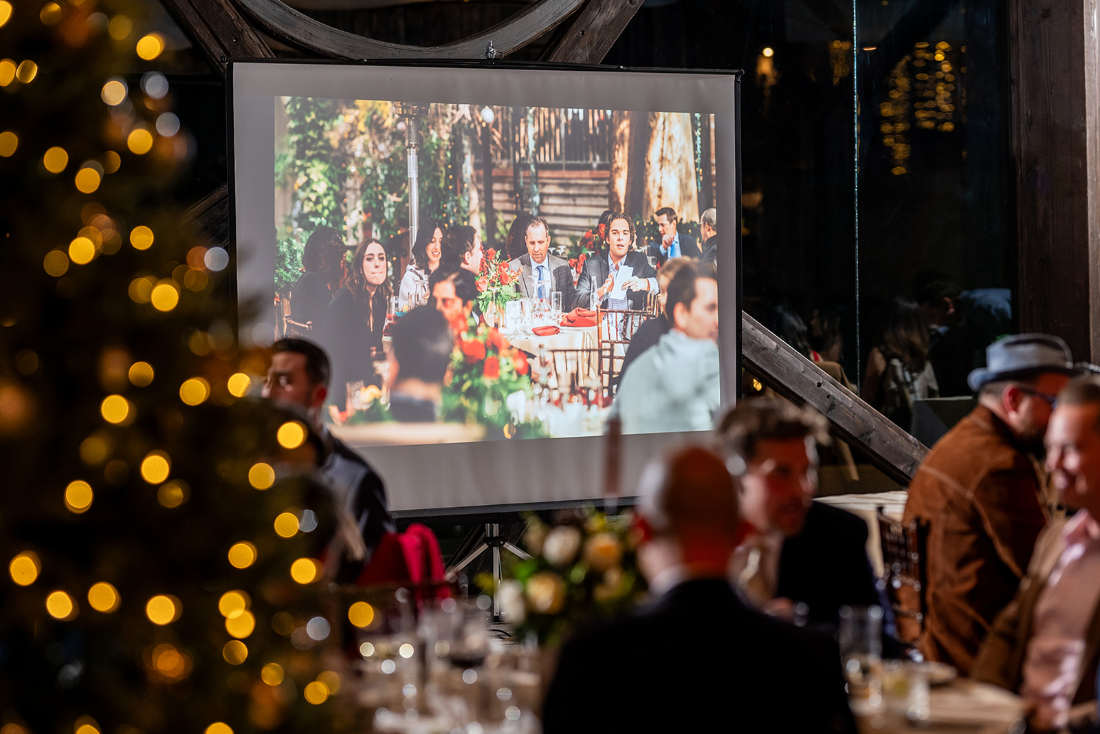 Large projection screen displaying event photography while guests dine at tables in a warmly lit reception space.
