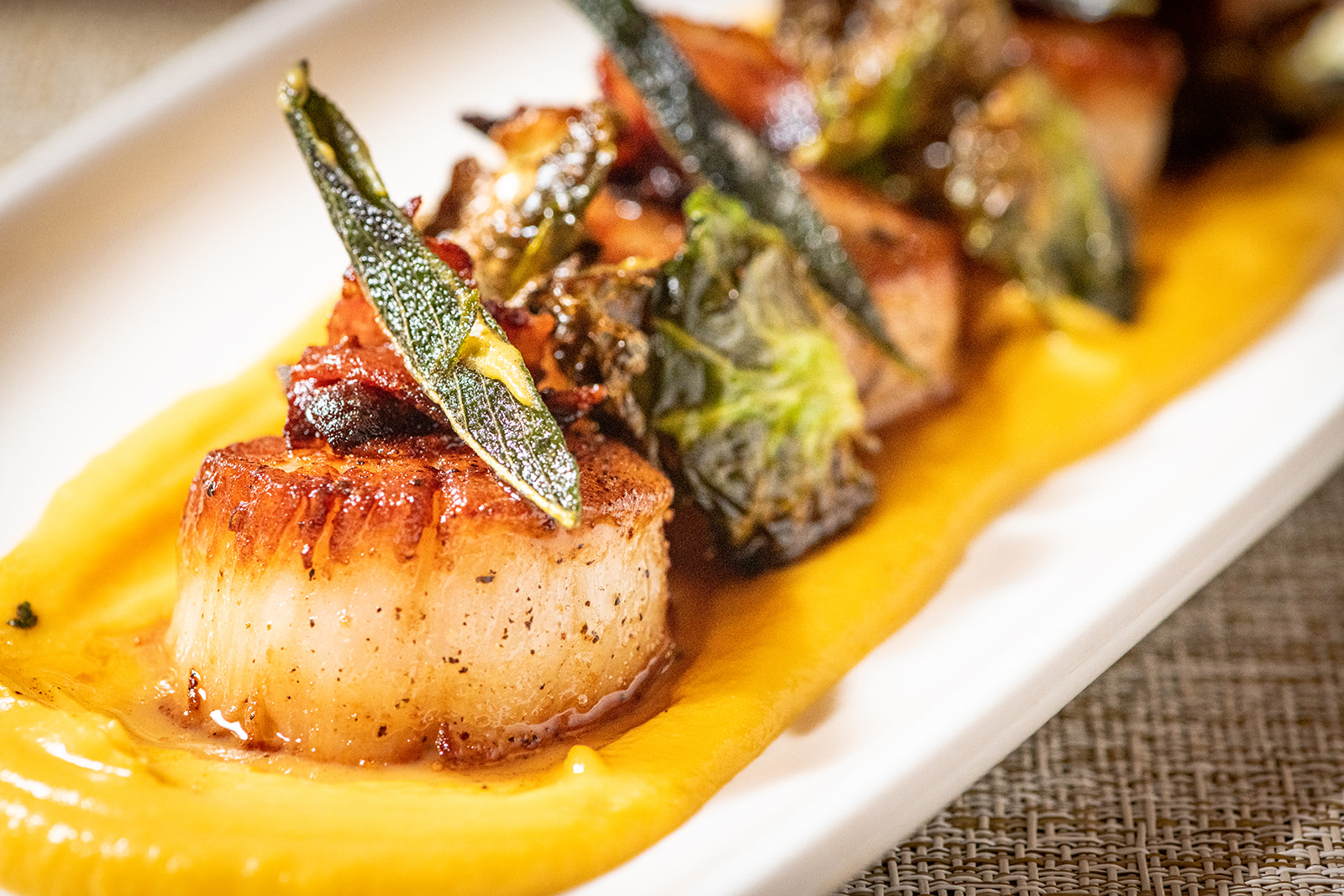 Close-up of a seared scallop plated with vegetables, herbs, and a bright yellow sauce.
