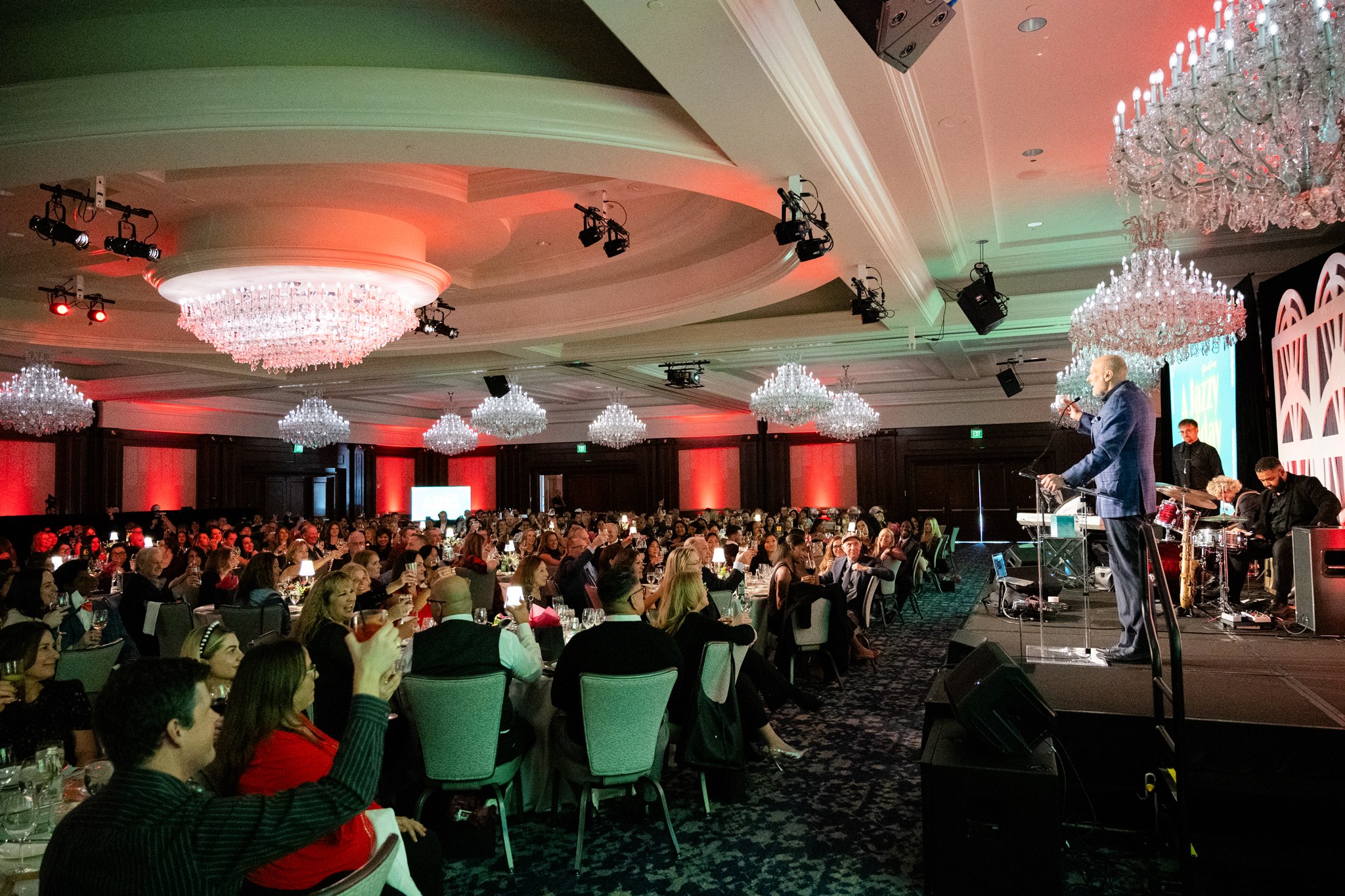 Large formal event in a chandelier-filled ballroom with a speaker addressing guests from a stage.