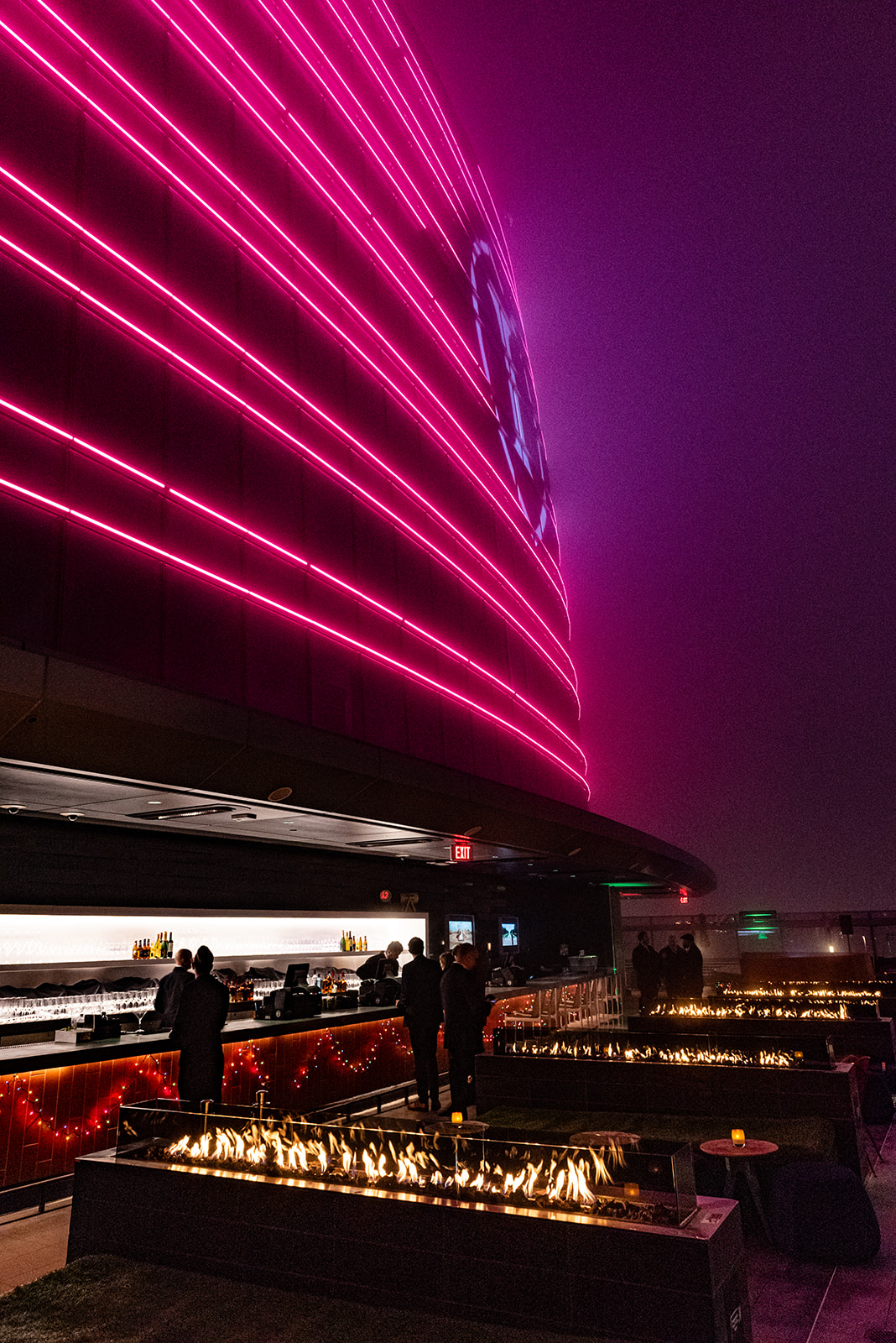 Modern outdoor event space at night with bright pink architectural lighting, fire features, and guests gathered near a bar.