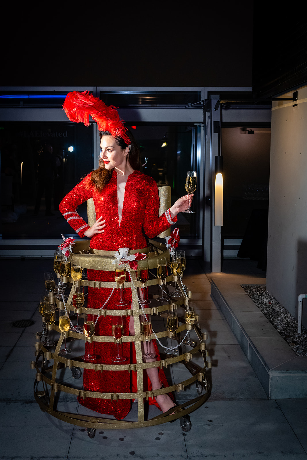Woman in a red sequined costume and feathered headpiece serving champagne from a wearable circular tray.