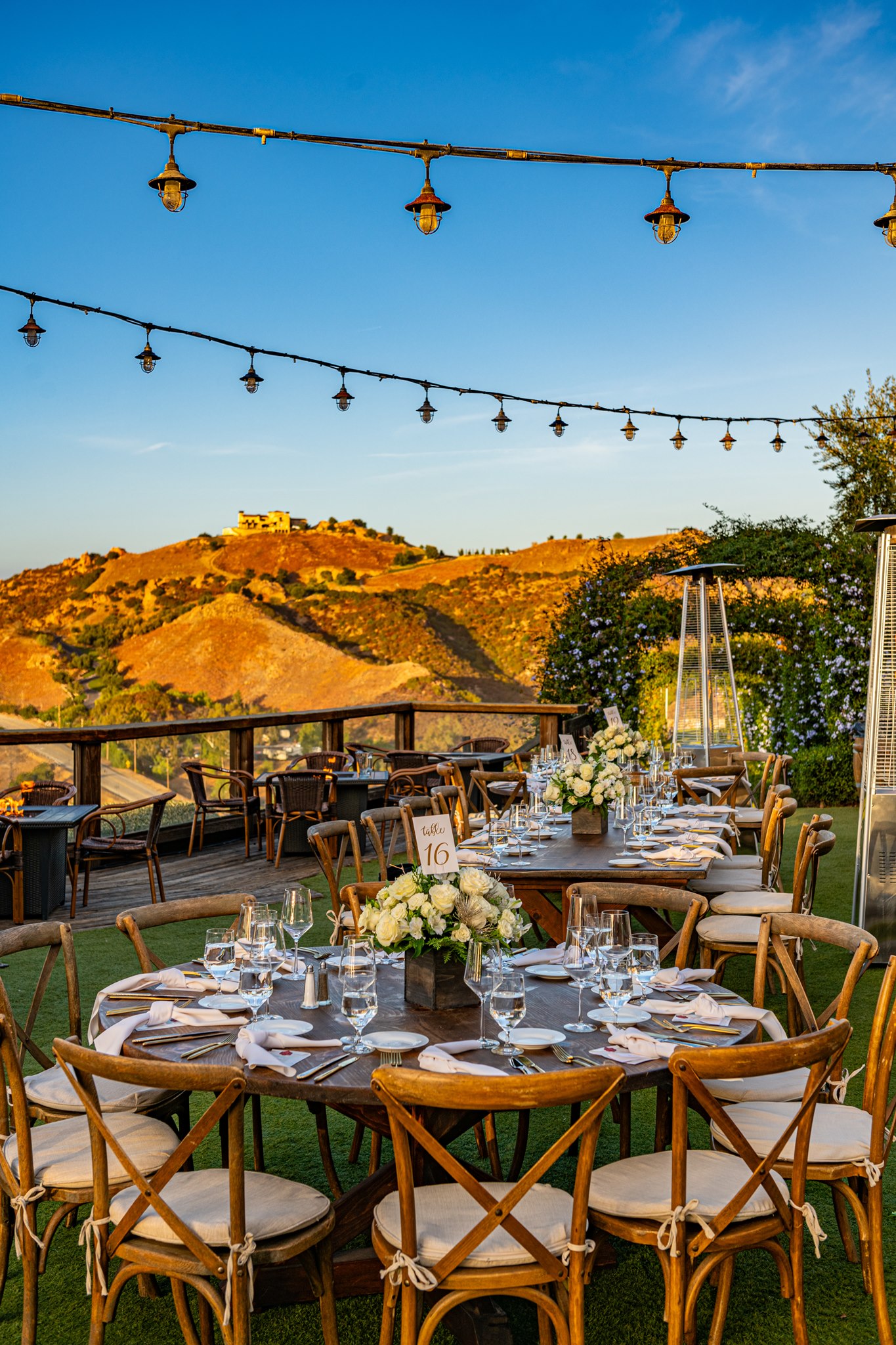 Outdoor reception tables with wooden chairs, white floral centerpieces, and string lights overlooking sunlit hills.