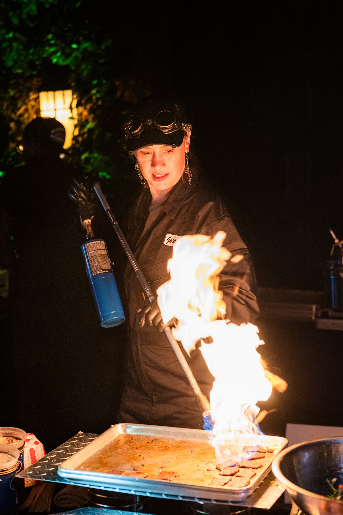 Chef using a blowtorch to sear food during a live outdoor cooking demonstration at night.