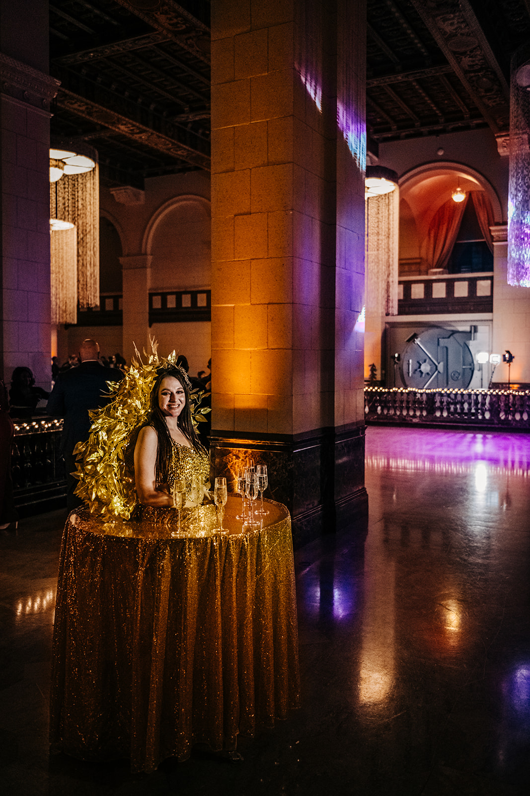 Woman in a gold sequined dress posing beside a champagne table at an elegant evening event with warm and purple lighting.