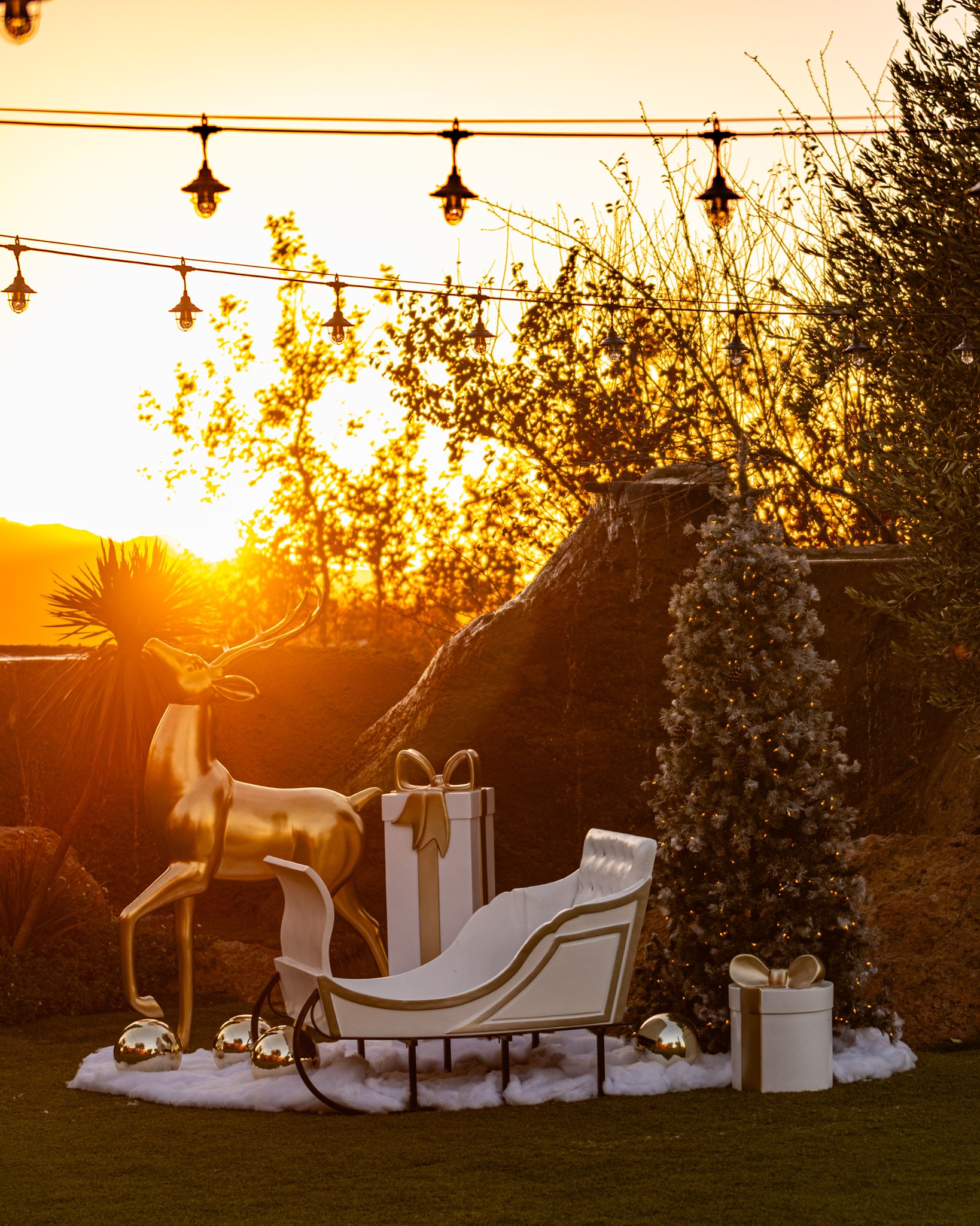 Outdoor holiday display at sunset with a gold reindeer, white sleigh, wrapped presents, string lights, and a lit Christmas tree.