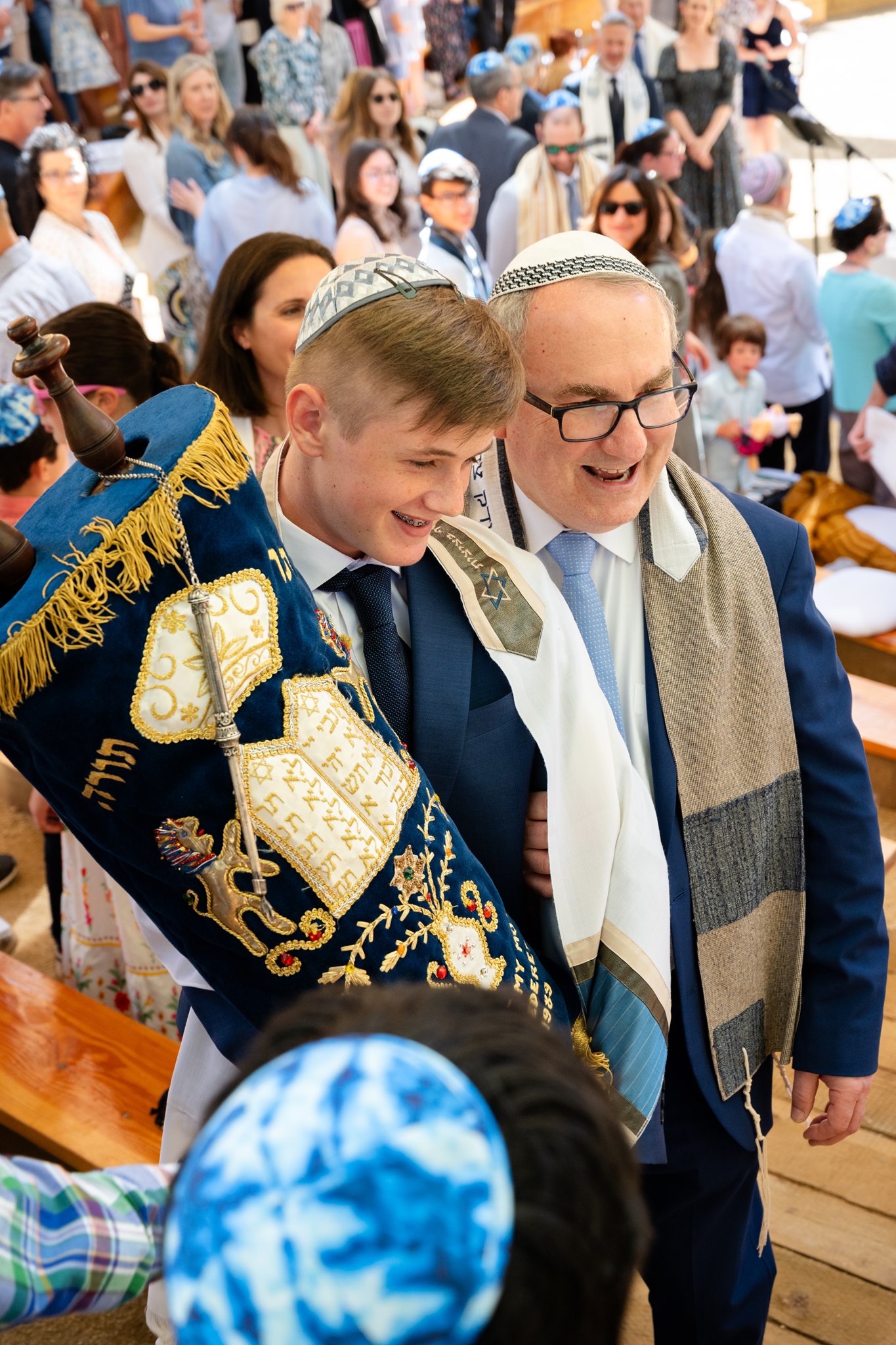 Teen holding a decorated Torah beside an older man wearing a prayer shawl during a crowded religious celebration.