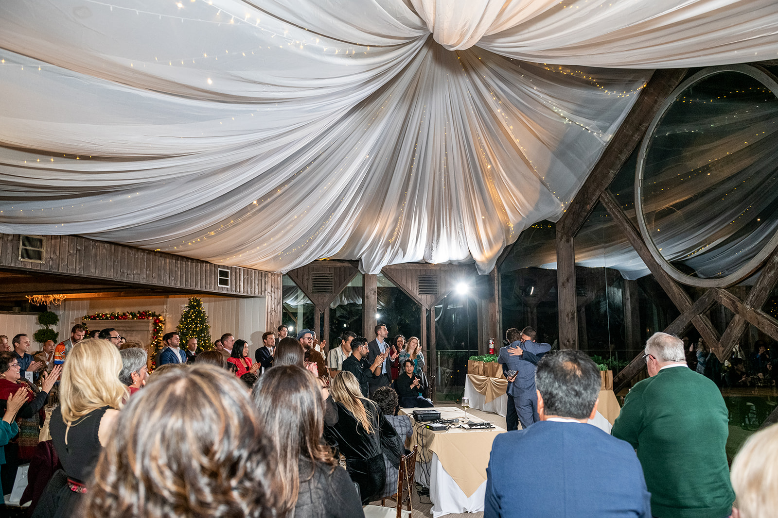 Guests gathered beneath a draped white ceiling and string lights while two people embrace during an indoor celebration.