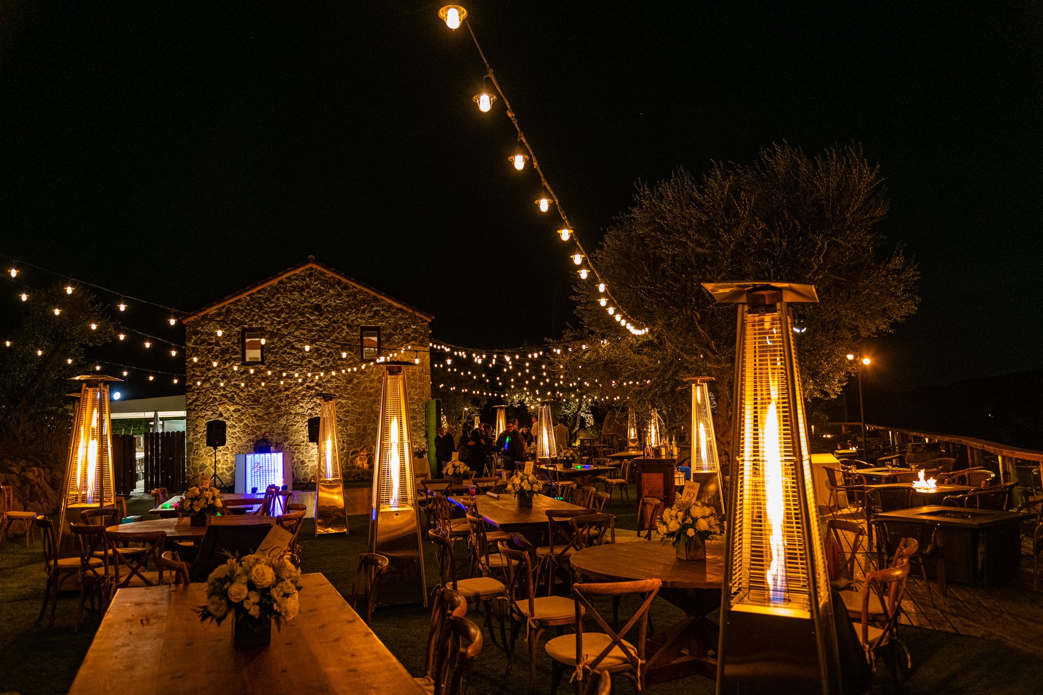 Outdoor evening reception with wooden tables, string lights, glowing patio heaters, and a rustic stone building.