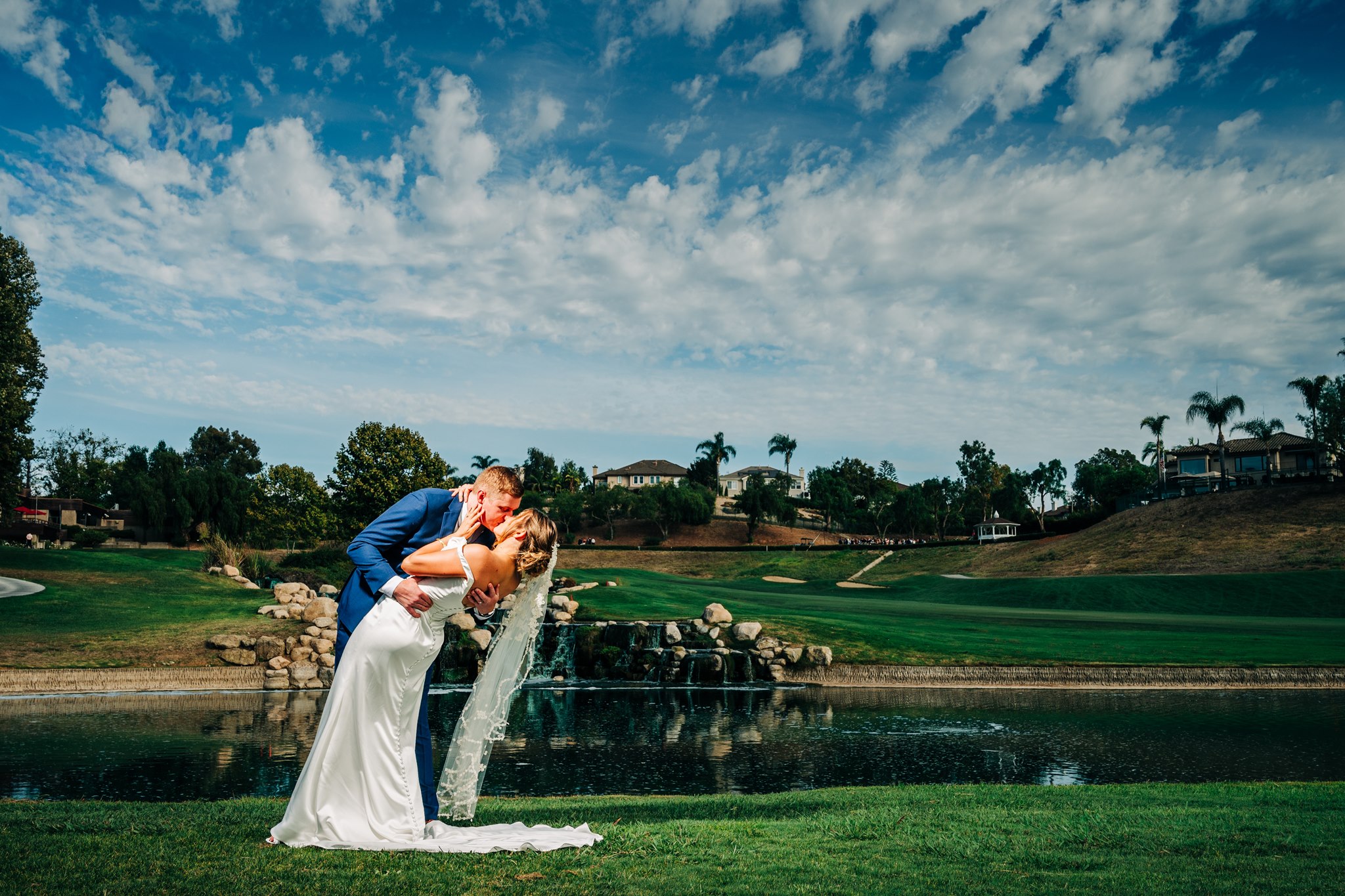 Bride and groom kissing beside a pond and waterfall on a landscaped golf course beneath a dramatic blue sky.