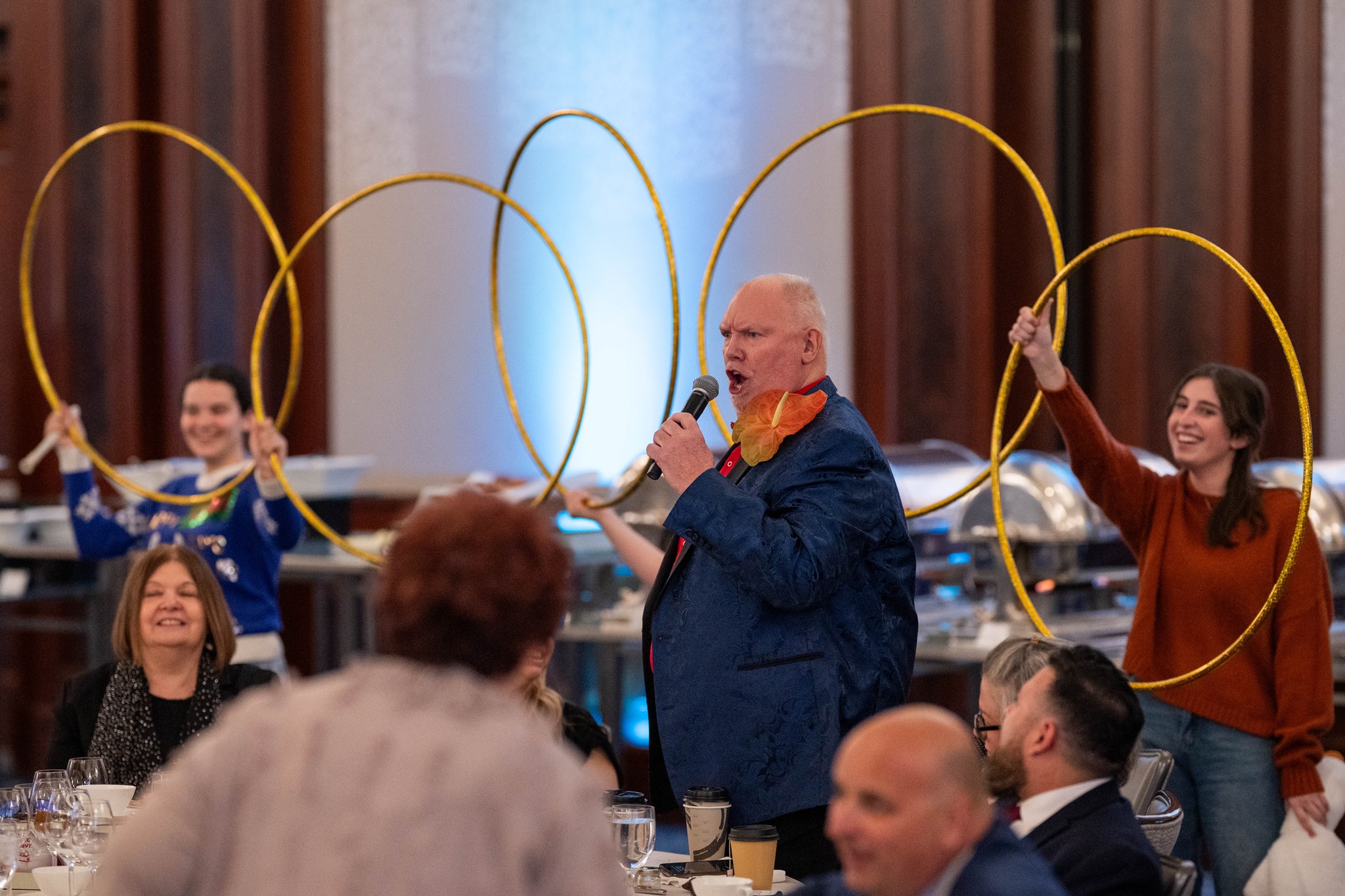 Entertainer speaking into a microphone while assistants hold large gold hoops among seated event guests.