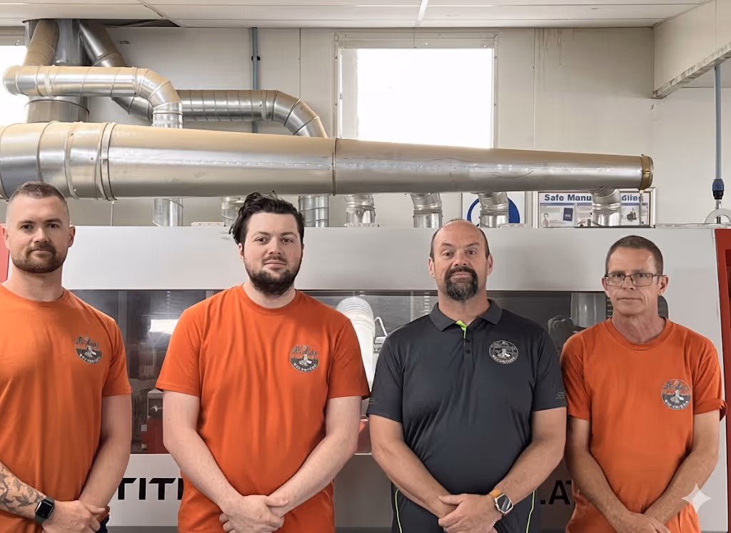 Four men standing indoors in front of industrial equipment, three wearing matching orange shirts and one wearing a black shirt.
