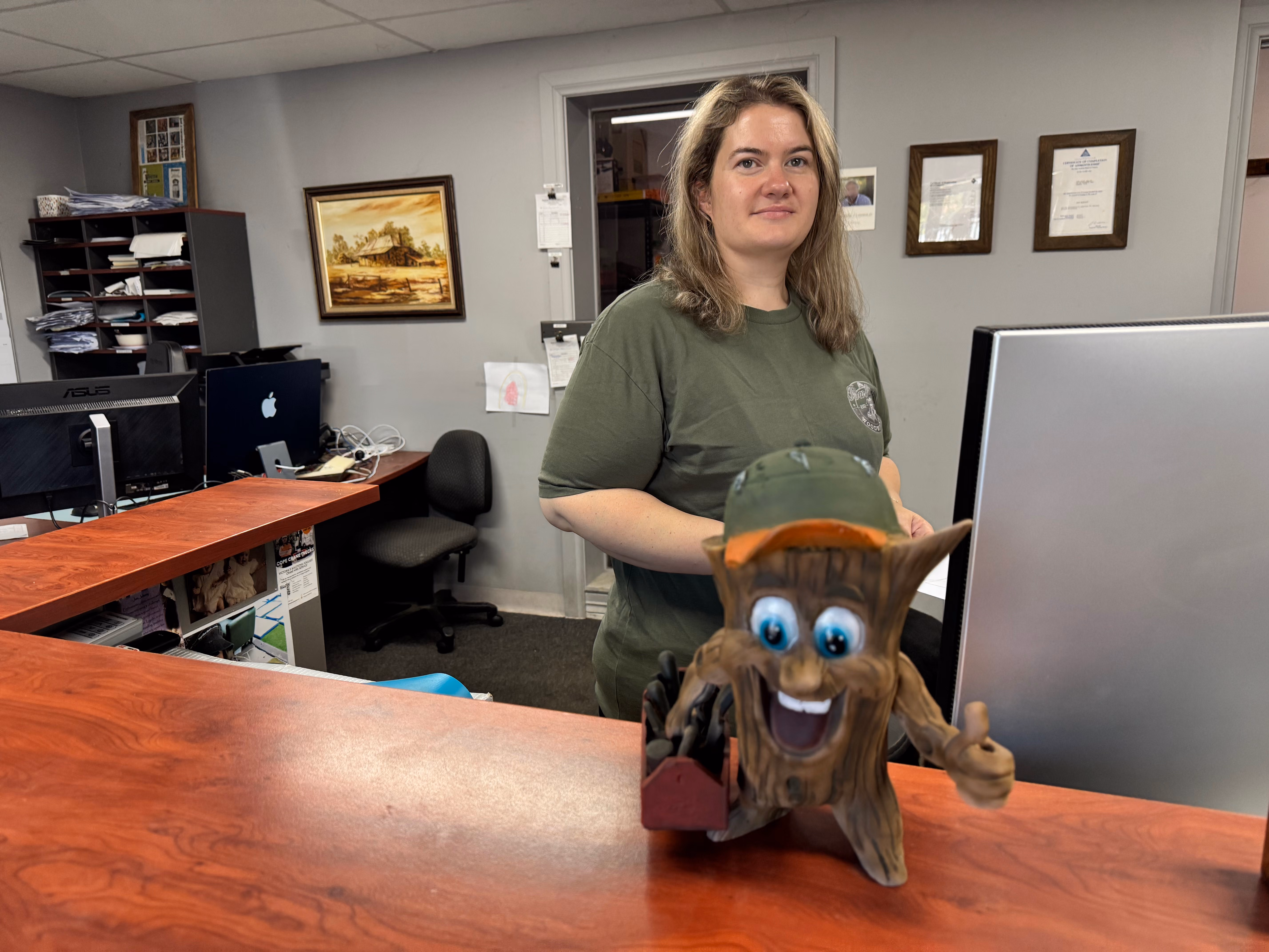 Woman with long hair wearing green shirt stands behind counter with smiling tree stump figure in front.