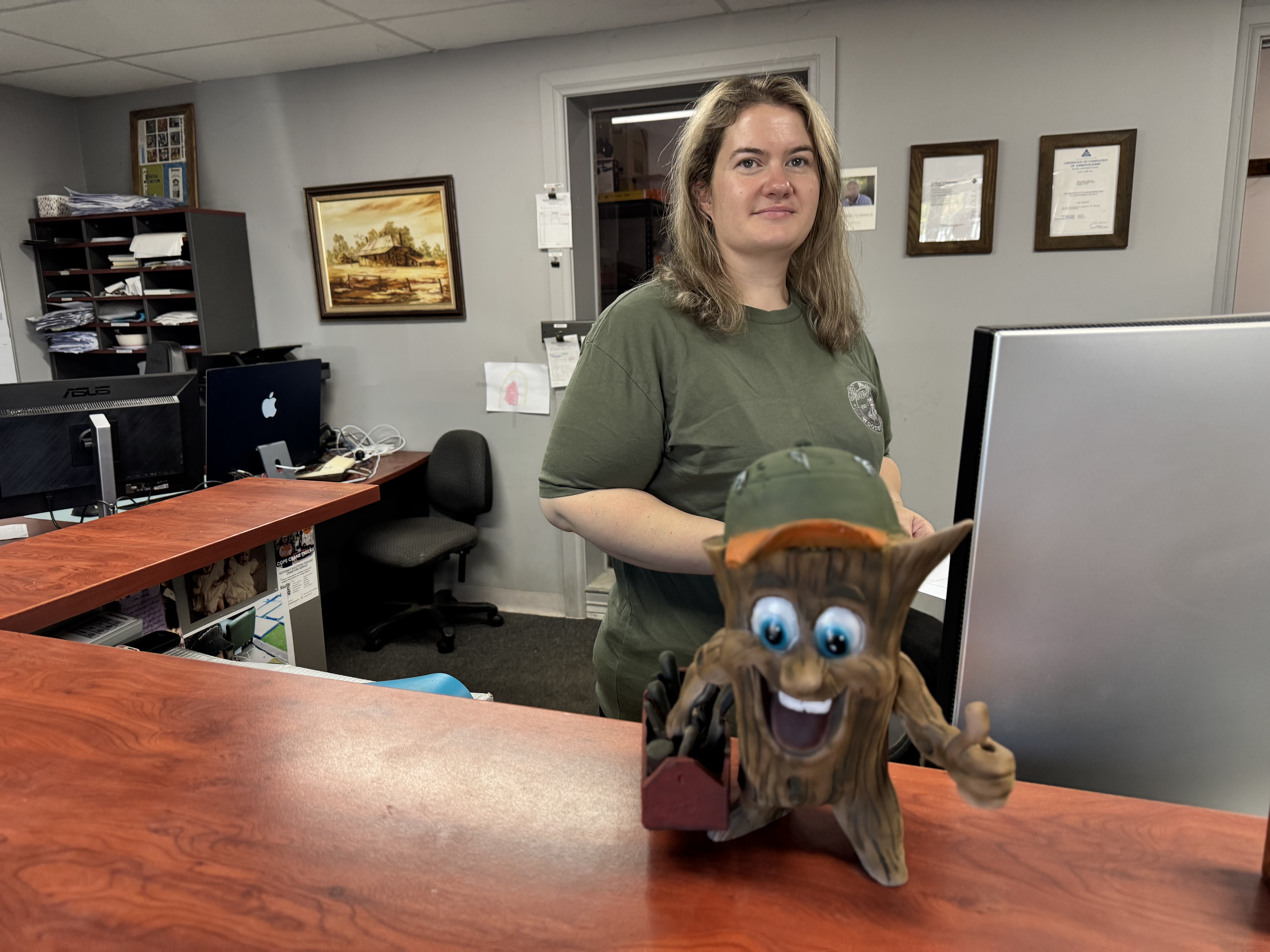 Woman with long hair wearing green shirt stands behind counter with smiling tree stump figure in front.