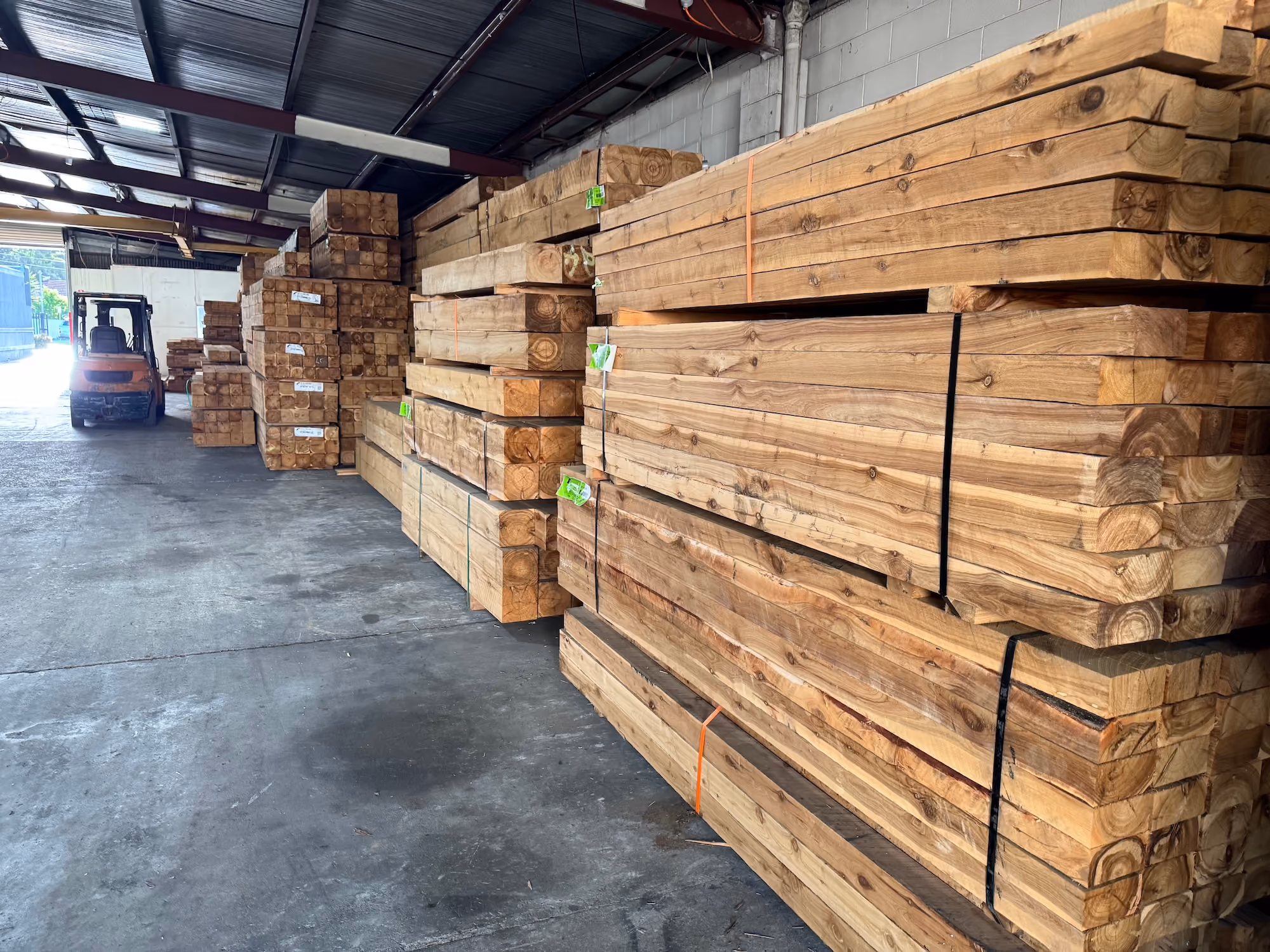 Stacks of large wooden beams stored inside a warehouse with a forklift at the back.