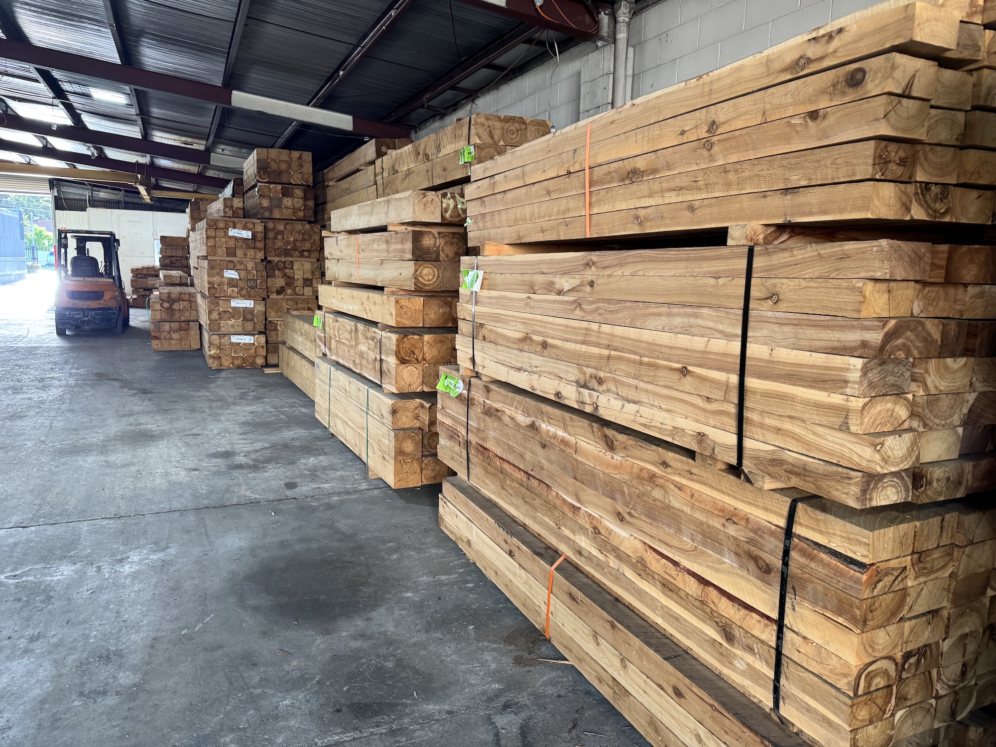 Stacks of large wooden beams stored inside a warehouse with a forklift at the back.