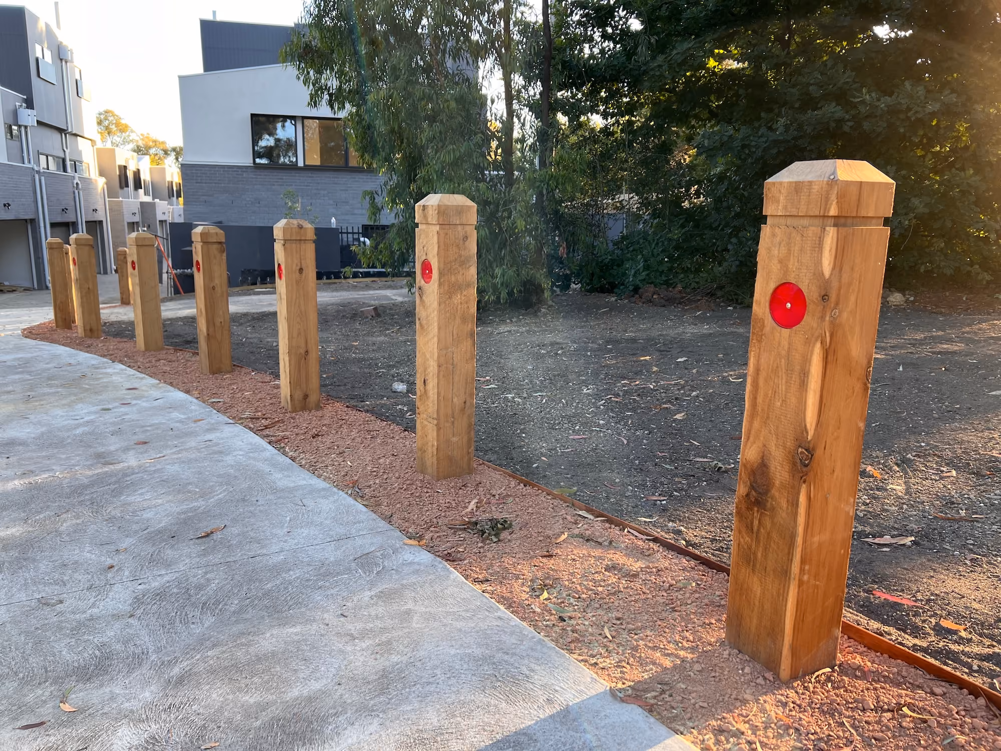 Row of wooden bollards with red reflectors along a curved concrete pathway next to a patch of bare soil and some trees, with modern buildings in the background.