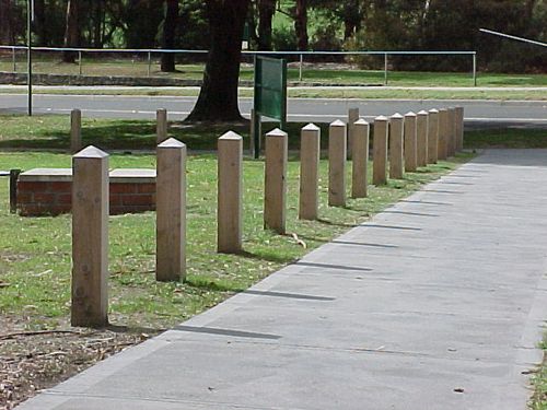 A row of wooden posts with red reflectors lined along a gravel and concrete path near modern buildings.