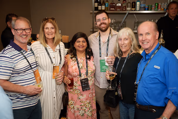 Group of 6 attendees with lanyards socializing at networking reception, holding drinks, bar shelves in background