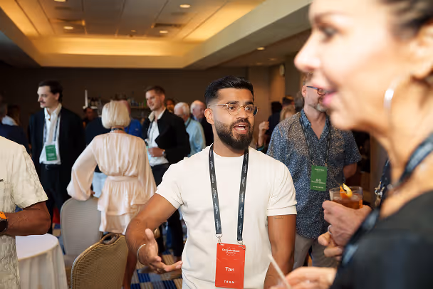 Man with red lanyard badge networking at event reception, gesturing animatedly, busy conference hall in background