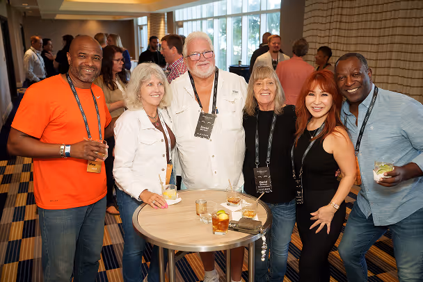 Group of 6 attendees smiling around a high-top table at networking reception, busy event hall in background