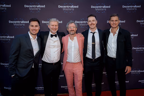 Five men posing on red carpet in front of Decentralized Masters branded backdrop at formal event