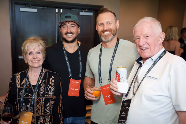 Four attendees posing together at networking reception, smiling with lanyards and drinks at event