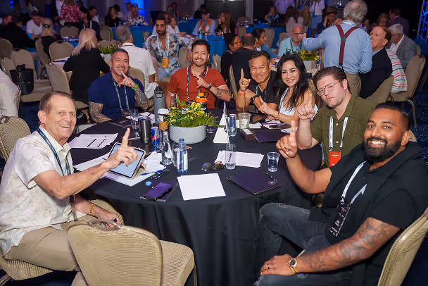 Group of 7 attendees giving thumbs up at conference table during event session, packed banquet hall in background