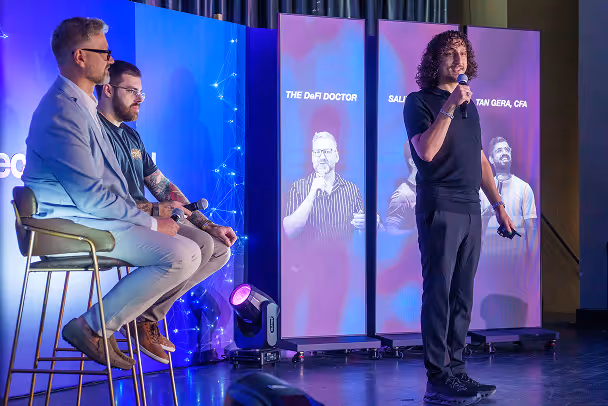 Speaker presenting on stage with microphone while two panelists sit on stools, LED screens showing speaker profiles in background