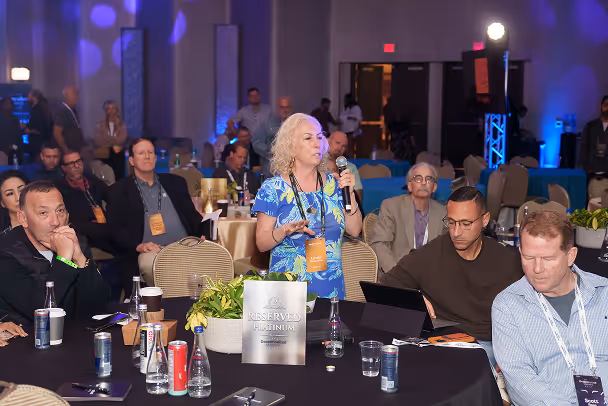 Woman standing with microphone speaking during conference session, attendees seated at table in blue-lit ballroom