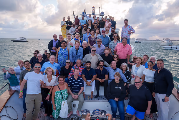 Large group photo of 50+ event attendees on yacht deck at sea with open water and boats in background