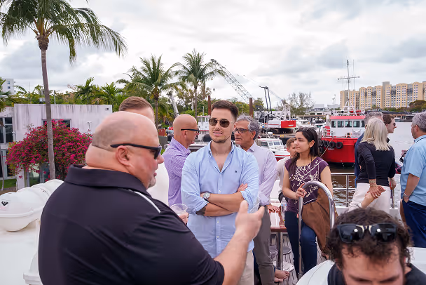 Attendees gathered at marina dock with palm trees and boats in background, socializing before yacht event