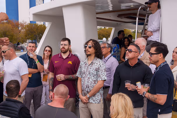 Group of attendees gathered on yacht deck listening at outdoor event, marina in background