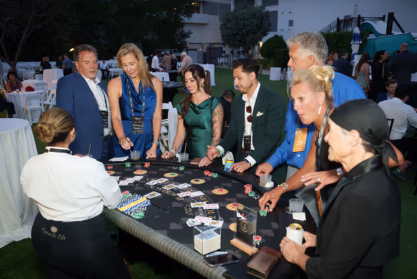 Two attendees in conversation at formal outdoor evening event with casino table setup in background