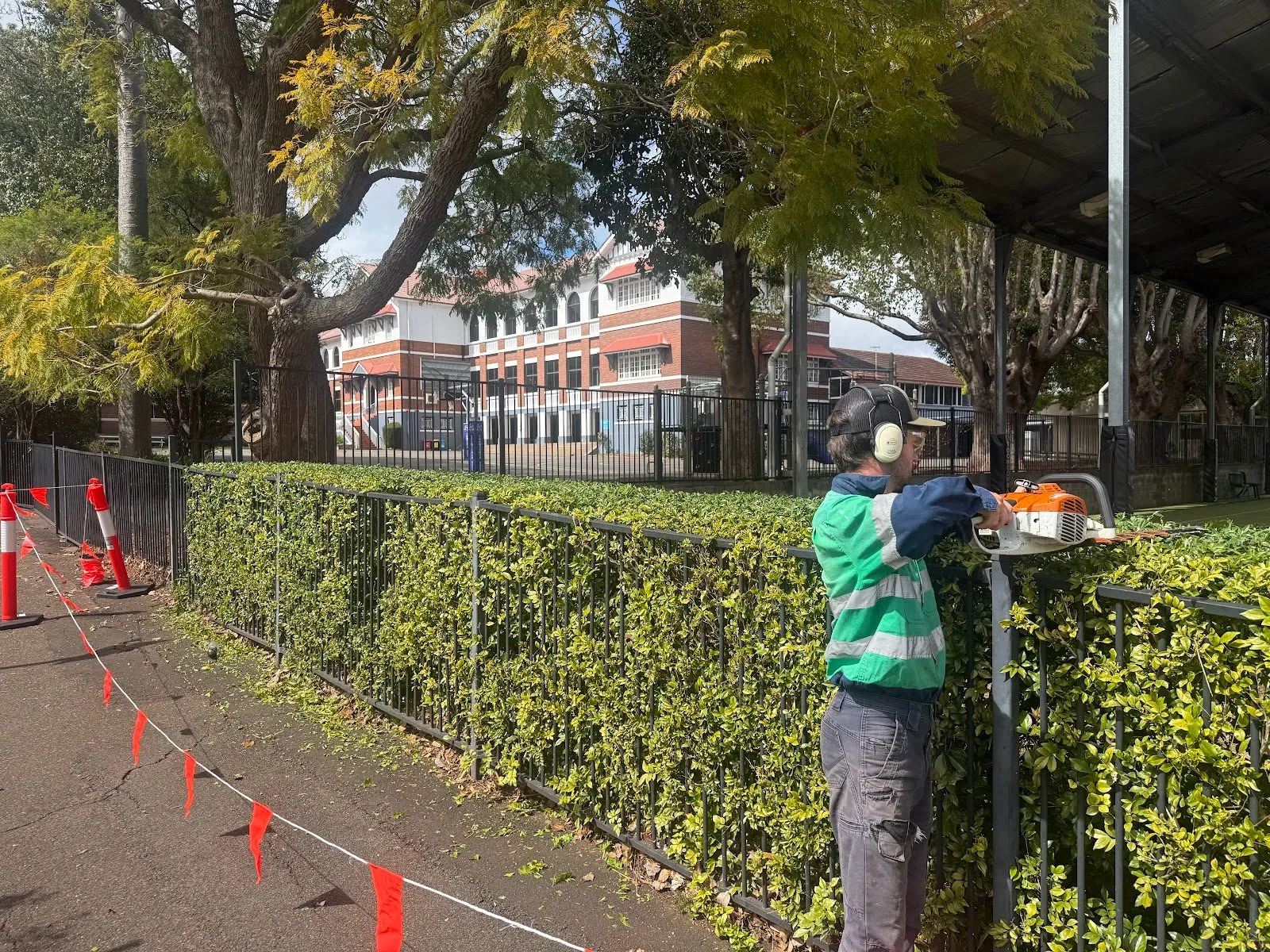 TopLawn crew member hedge trimming along a school fence line, work area taped off