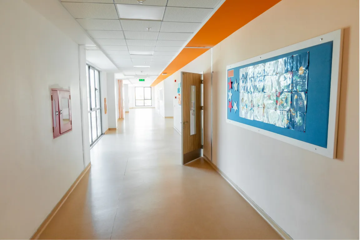 Leerer Schulflur mit einer blauen Pinnwand voller Kinderbilder und einem orangefarbenen Deckenband