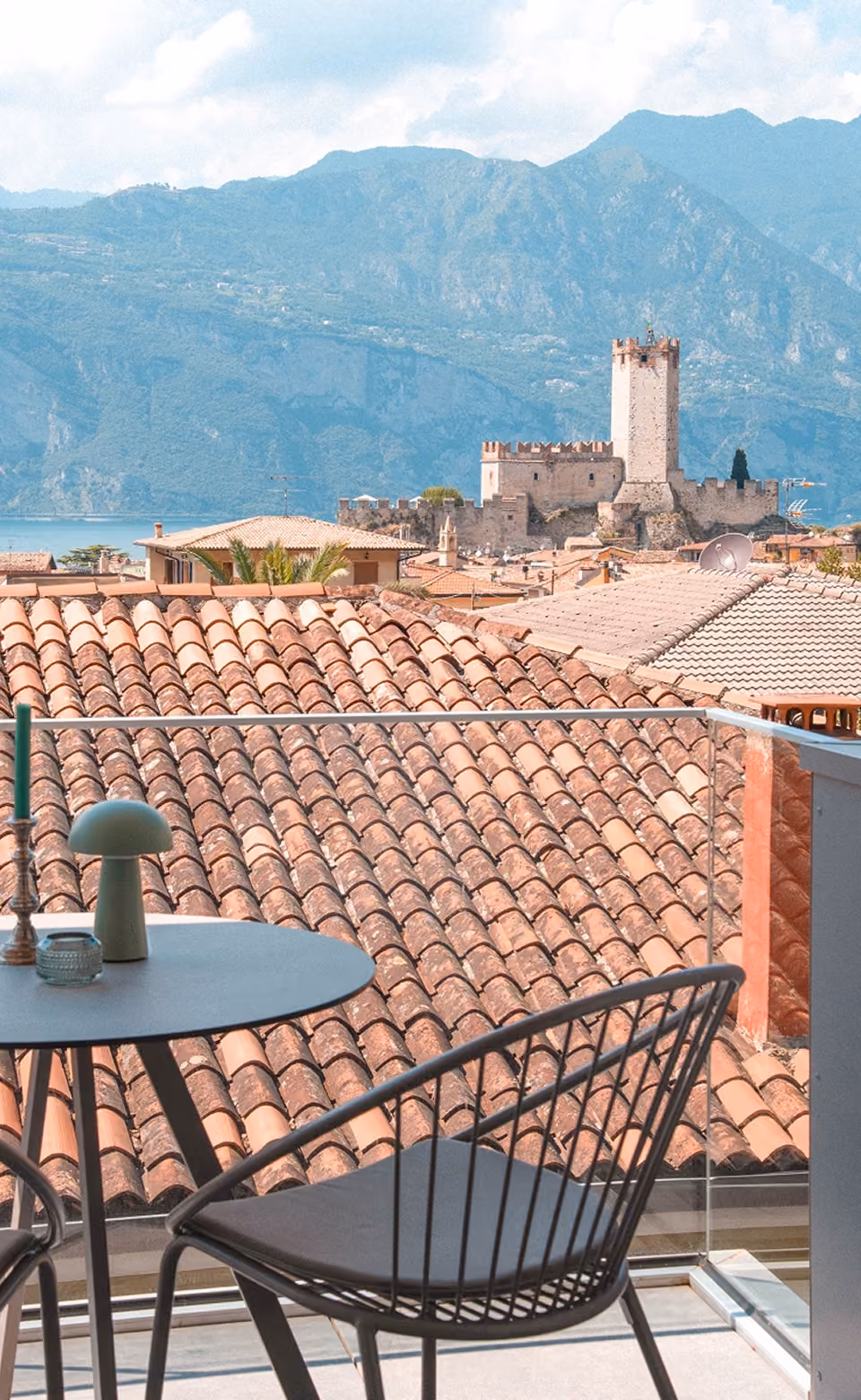 Balcony with table and chairs overlooking terracotta roofs, historic castle, lake, and mountains in the background.