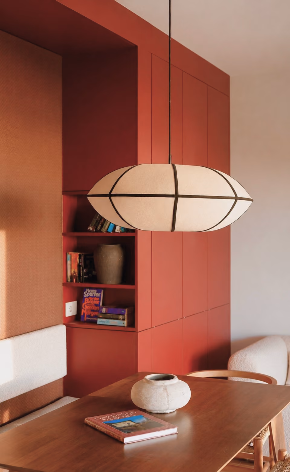 Modern dining area with a wooden table, white vase, hanging lamp, and red built-in shelves with books.