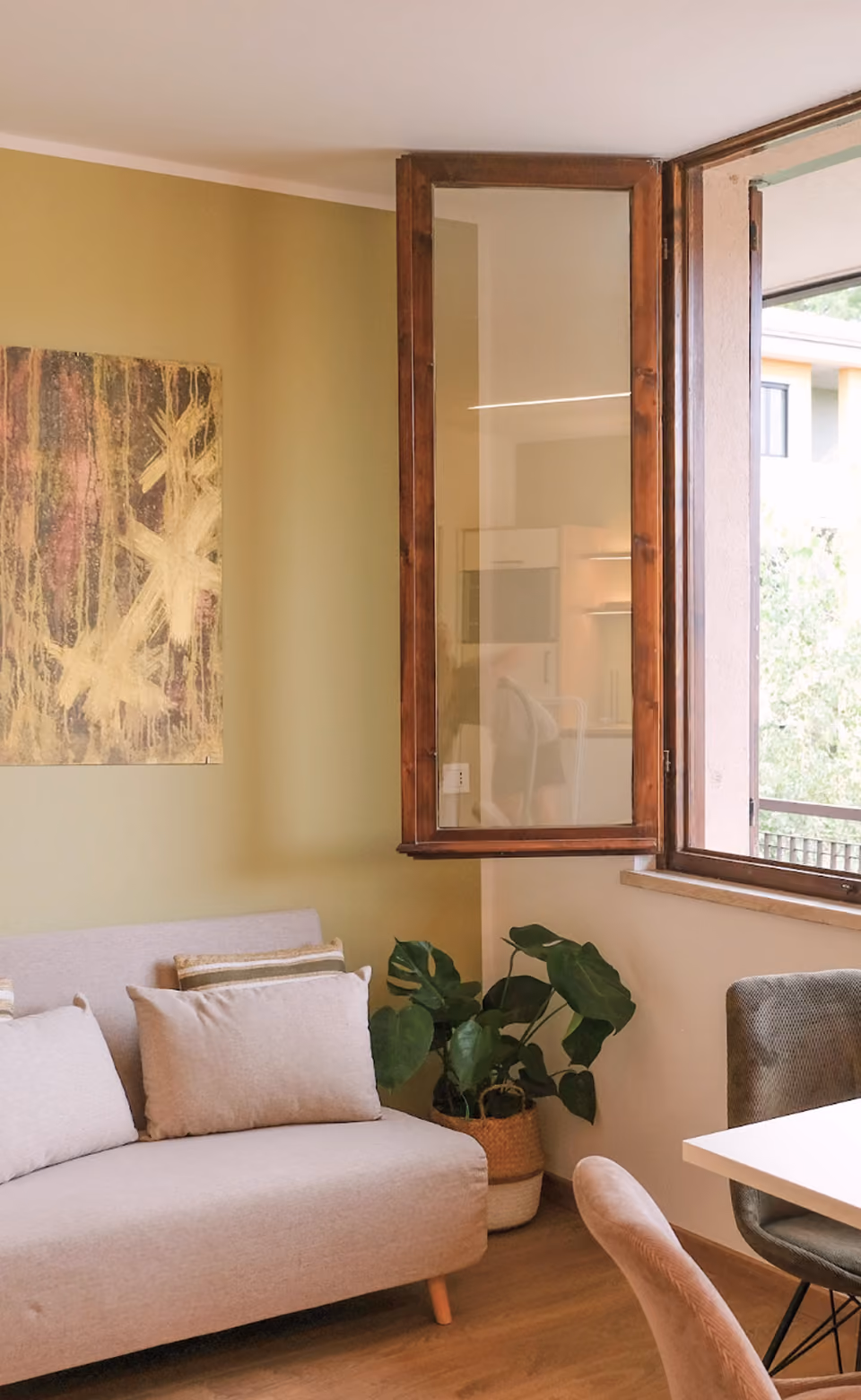 Cozy living room corner with beige sofa, green potted plant, open wooden window, and abstract wall art.